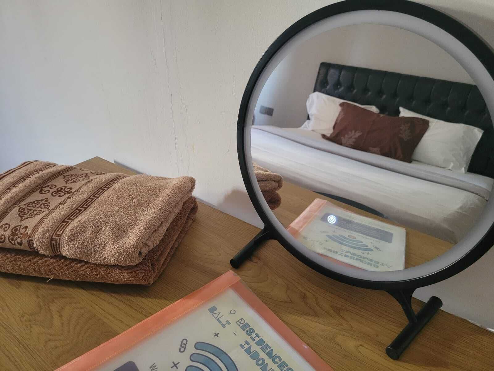 A neatly folded brown towel on a wooden table, a round mirror reflecting a bed with white and brown bedding, and a visible hotel room number sign.