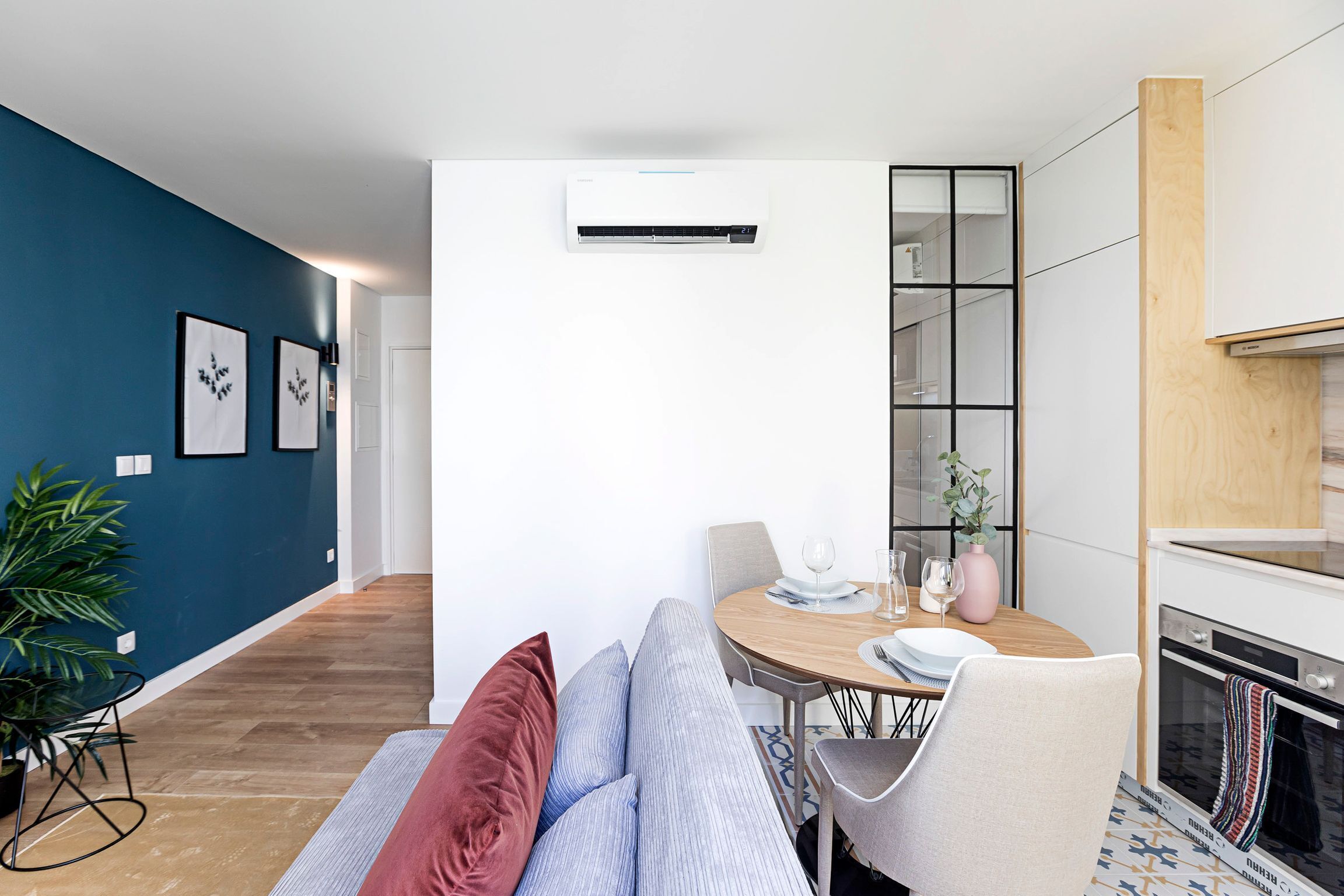 Ein modernes Wohnzimmer mit einer Couch, einem Esstisch mit Stühlen, einer Küchenzeile mit Herd und einem großen Fenster.
