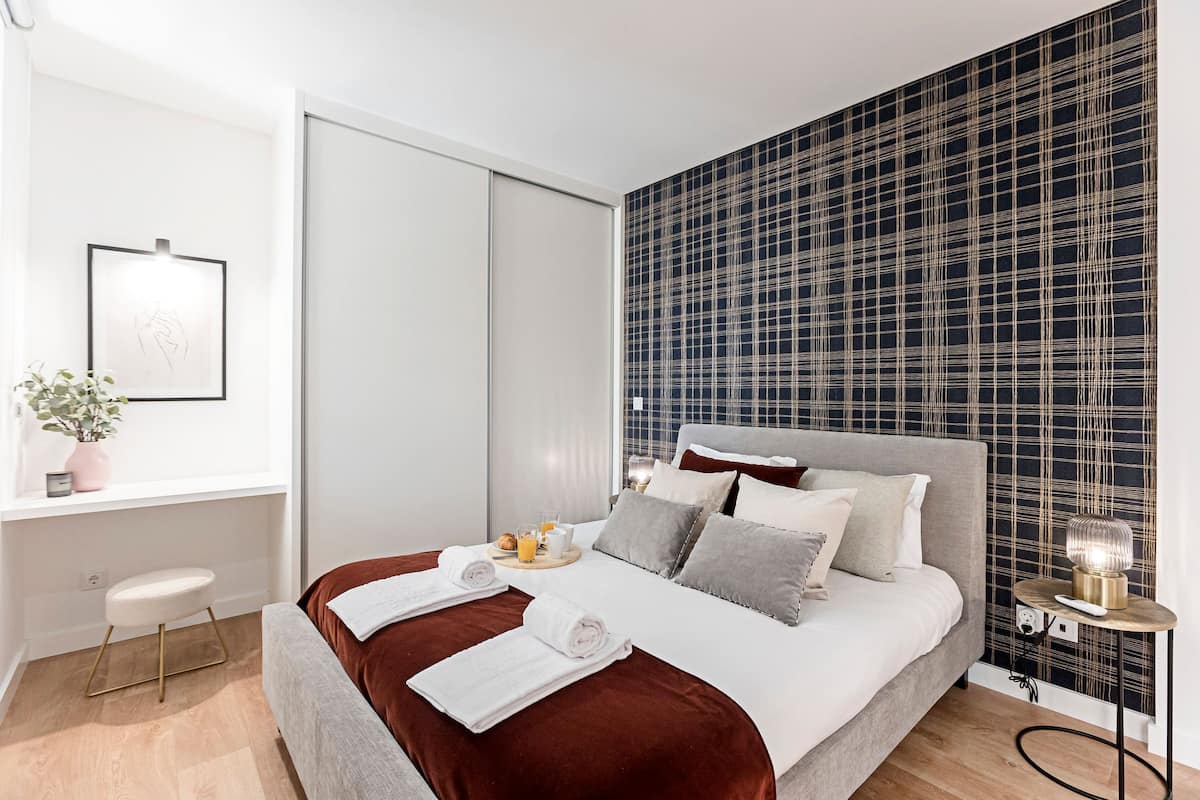 A modern bedroom with a large bed, a bedside table with a lamp, a small stool, and a window with a plant.