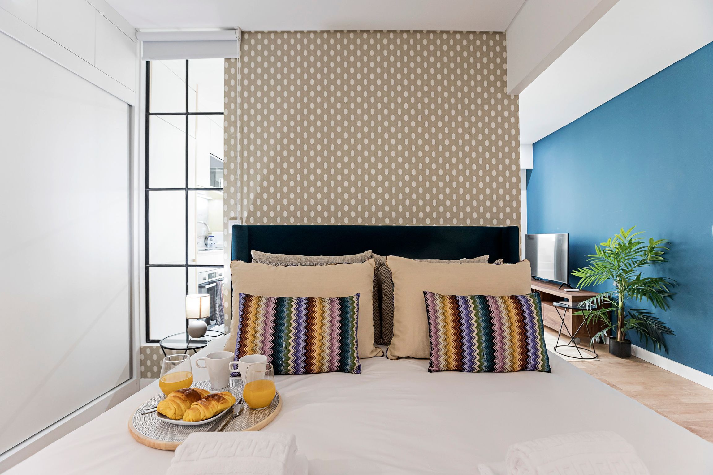 A modern hotel room with a large bed, colorful pillows, a breakfast tray, and a view of the city through large windows.