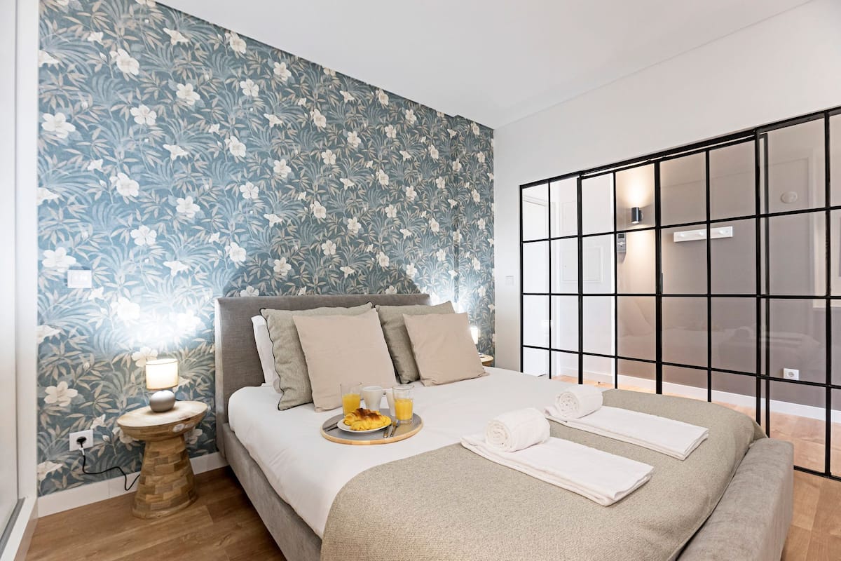A modern bedroom with a floral wallpaper, a bed with a grey headboard, and a bedside table with a lamp and breakfast items.
