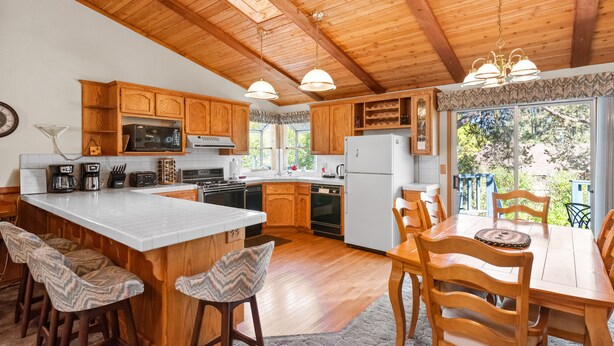 A kitchen with wooden cabinets, a large island with bar stools, a dining table with chairs, and a view of the outdoors through large windows.
