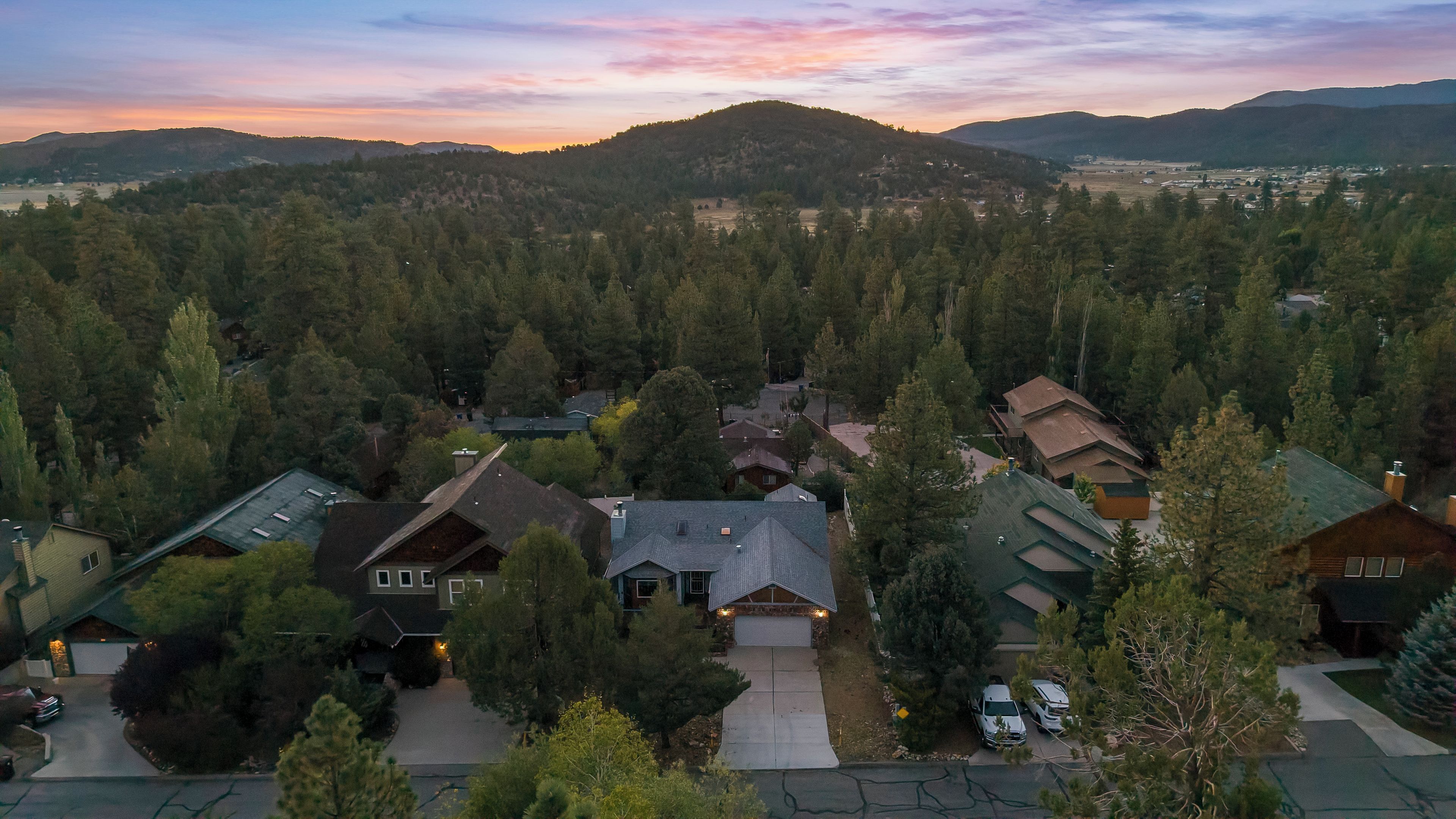Un quartier résidentiel où les maisons sont entourées d’une forêt dense, sous un ciel dégagé, avec des montagnes qui se dessinent à l’horizon.