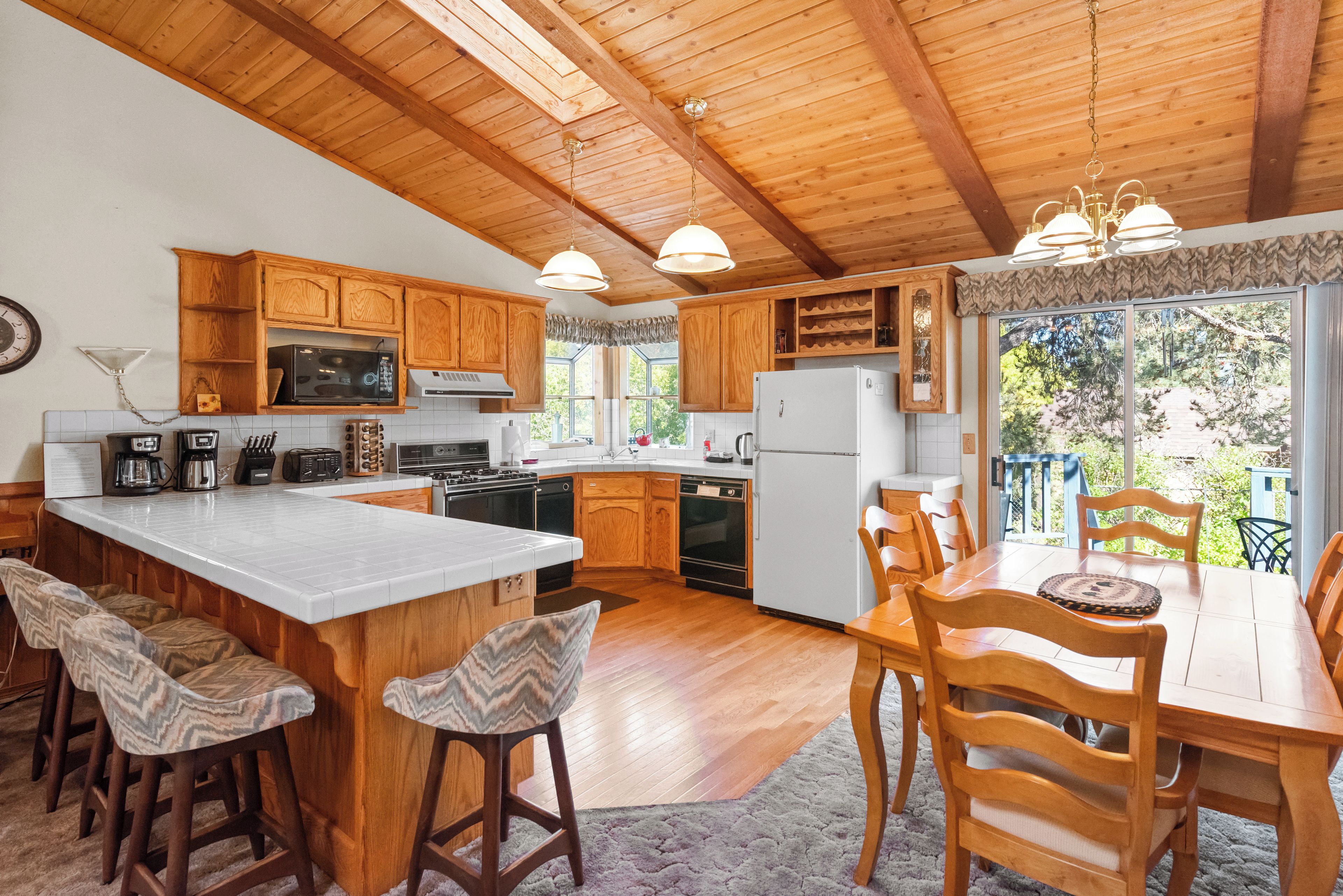 Une cuisine avec des meubles en bois, un grand îlot central doté de tabourets, une table à manger avec des chaises, et une vue sur l’extérieur grâce à de grandes fenêtres.