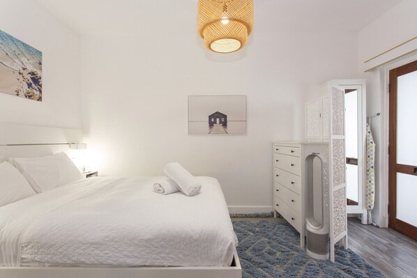 A bedroom with a bed, a white dresser, a wall-mounted artwork, a ceiling light fixture, and a door leading to another room.