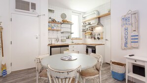 A kitchen with white cabinets, a round dining table with chairs, a clock, and a wall-mounted shelf with kitchen utensils. - Beach Shack Fremantle (Fremantle)