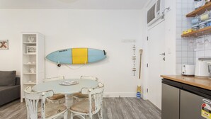 A kitchen with a surfboard, a dining table with chairs, a white shelf, and a grey sofa. - Beach Shack Fremantle (Fremantle)