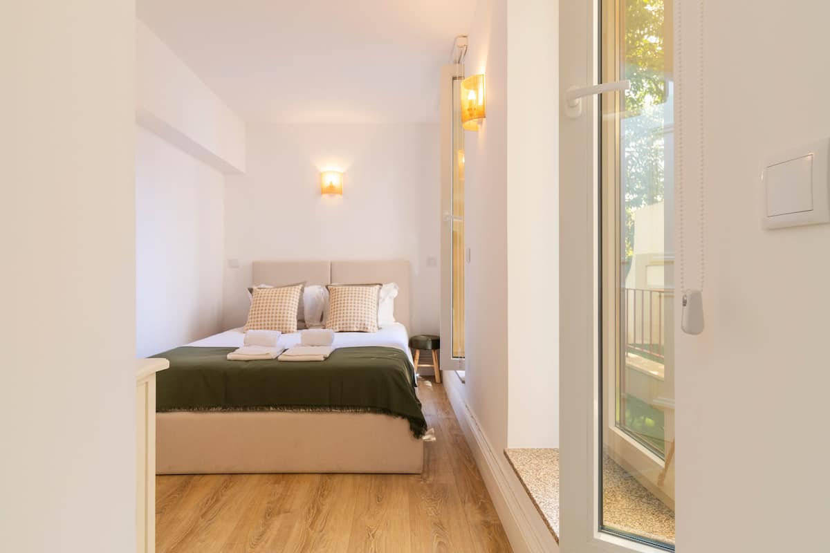 A bedroom with a bed, pillows, a green blanket, and a nightstand. There is a sliding glass door leading to an outdoor area.