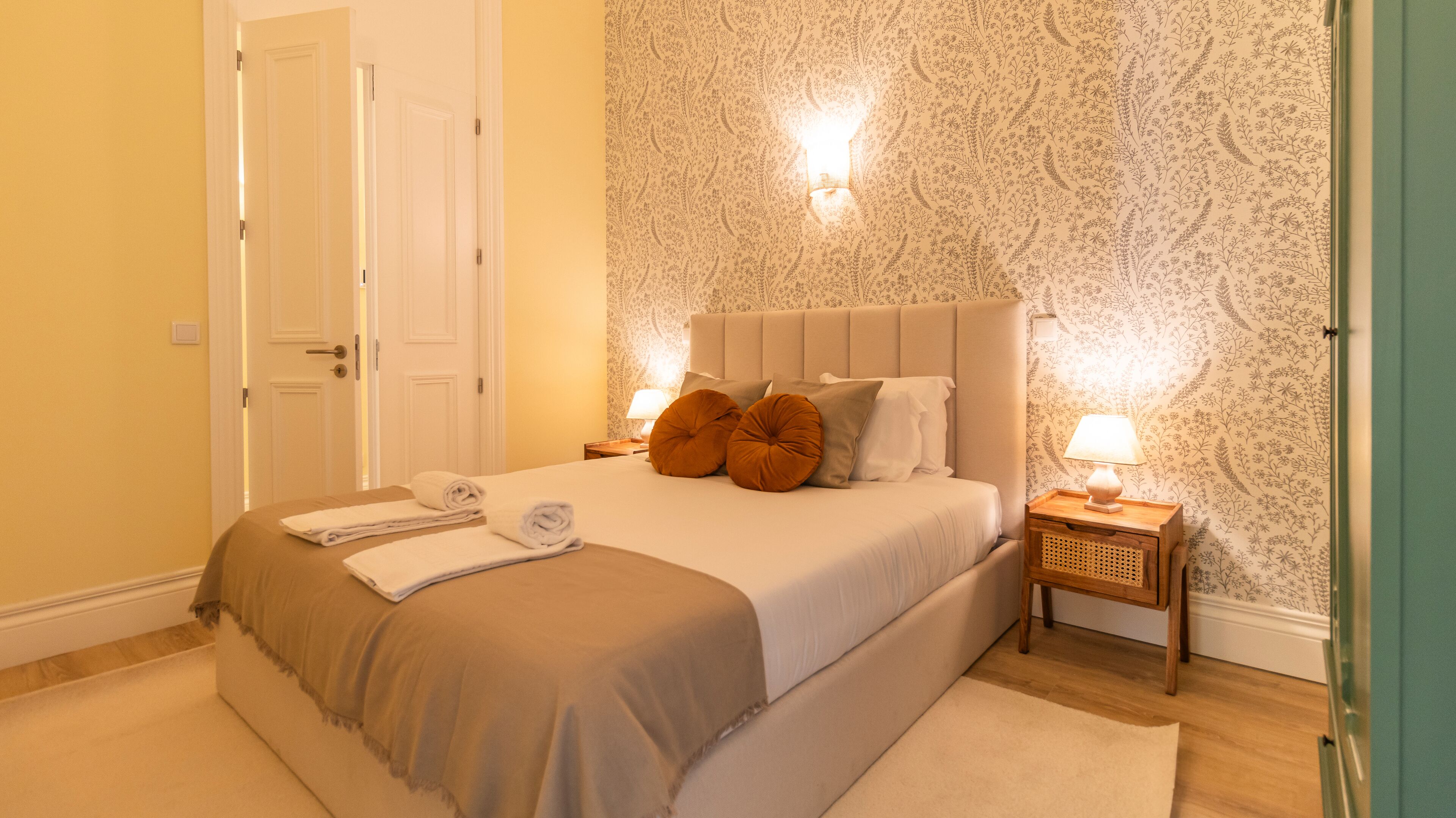 A neatly made bed with a beige headboard, two pillows, and a folded towel on the side. There is a bedside table with a lamp and a small wooden nightstand with a lamp. The room has floral wallpaper and a door to the left.