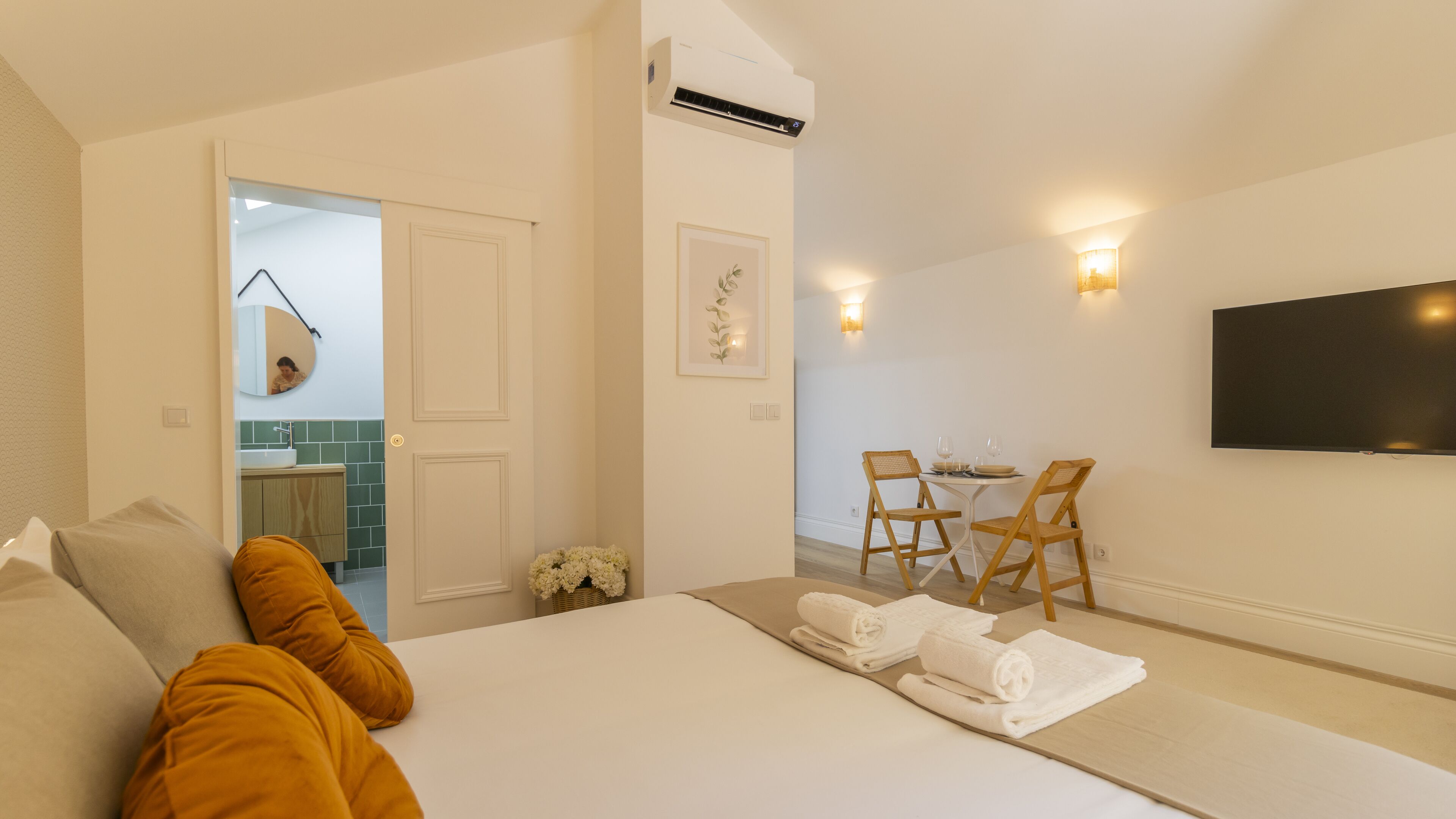 A bedroom with a bed, a TV, a dining area with chairs and a table, and an air conditioning unit.