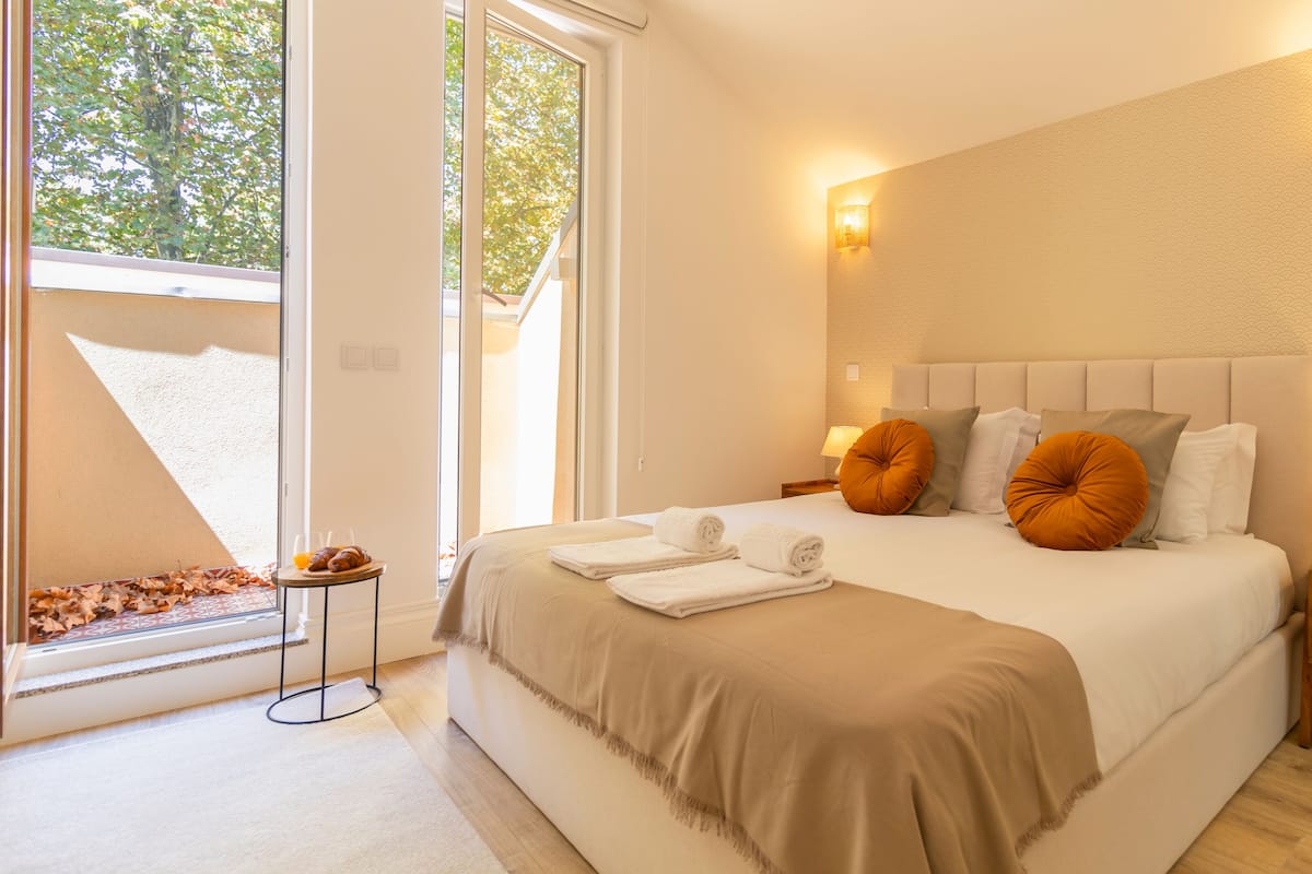 A hotel room with a large bed, a bedside table with a lamp, and a view of trees through the window.