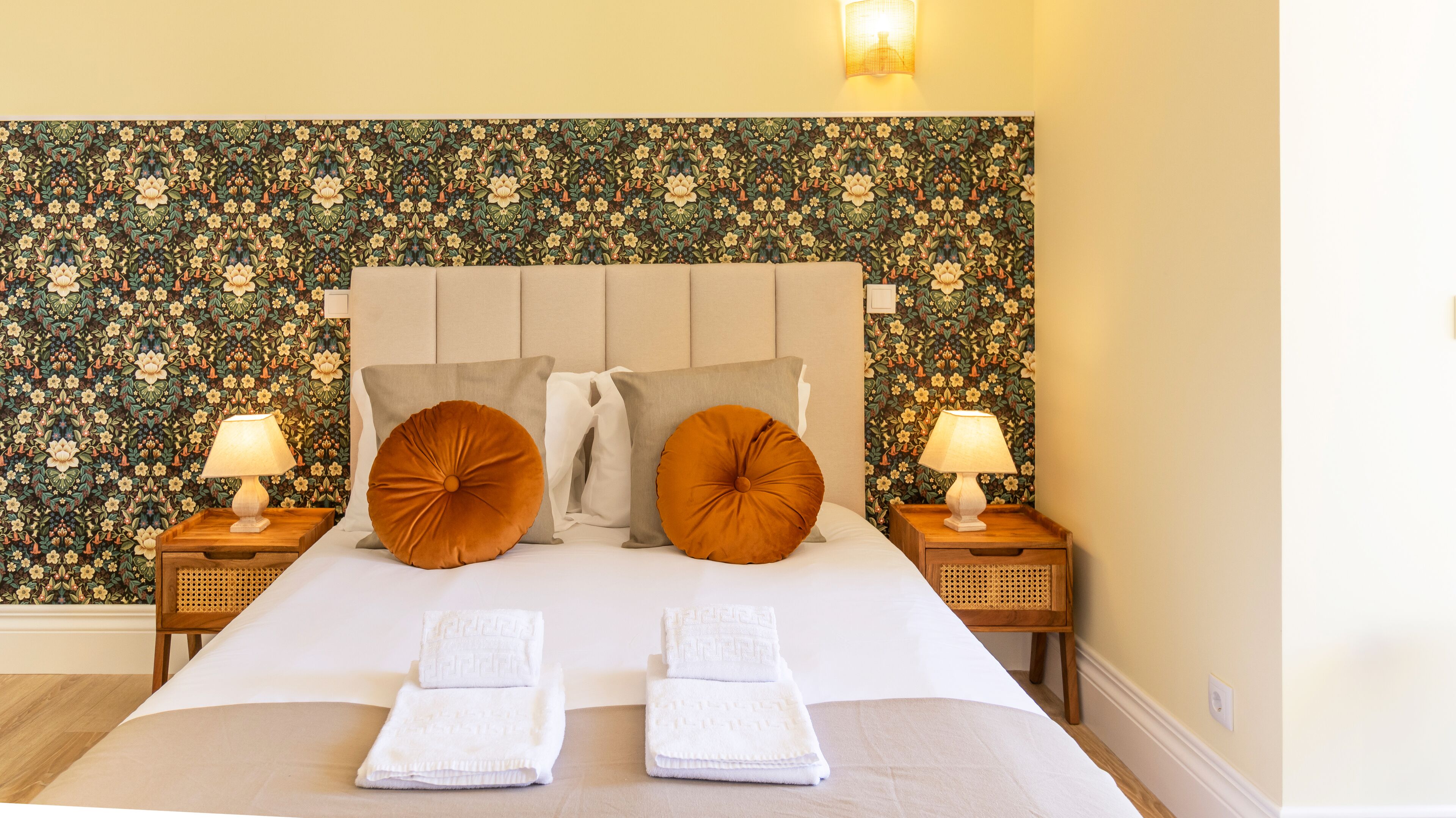 A neatly made bed with two pillows, two bedside tables with lamps, and a floral wallpaper.