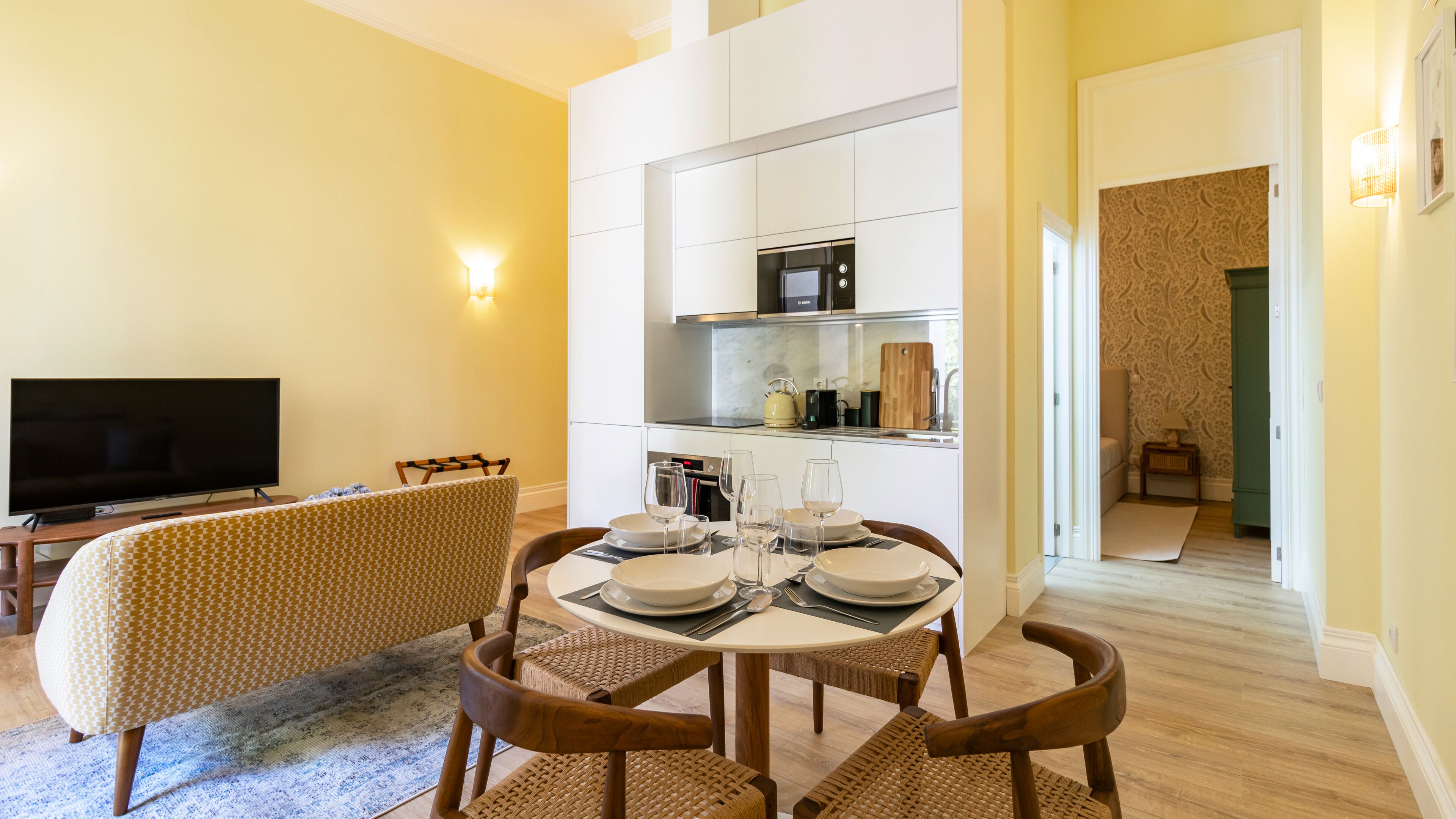 Una sala moderna con televisor de pantalla plana, un sofá, una mesa de comedor preparada para cuatro comensales y una zona de cocina con microondas y hervidor.