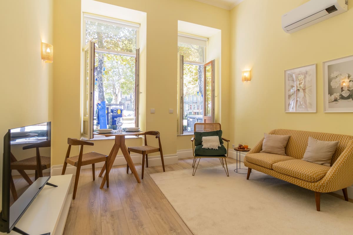 A living room with a sofa, a dining table with chairs, and two windows with views of the street.