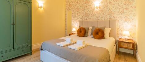 A hotel room with a bed, two nightstands, a green wardrobe, floral wallpaper, and a wooden floor.
