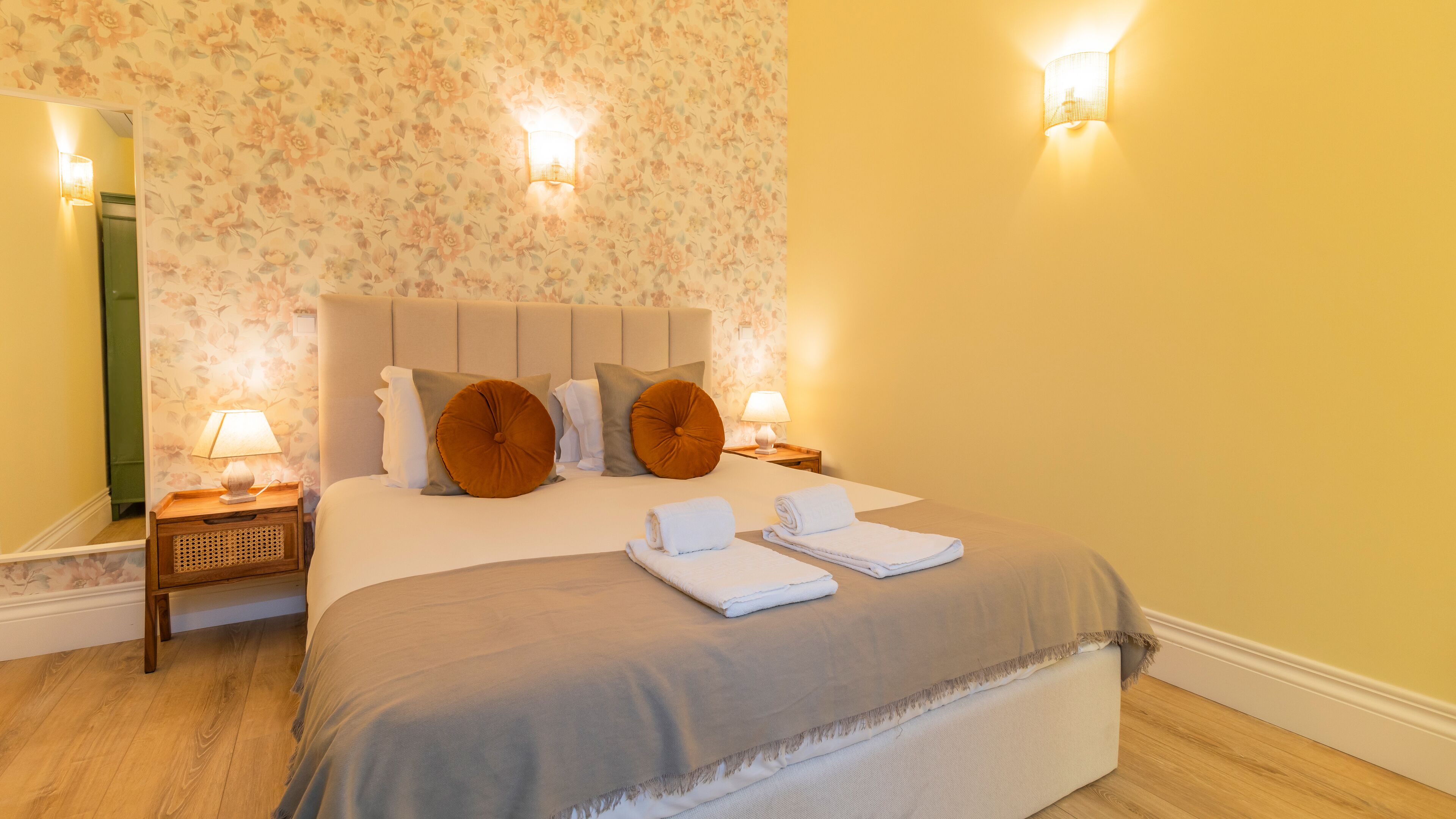 Un letto rifatto con ordine, con copriletto grigio, due asciugamani bianchi e due cuscini decorativi marroni. Sono presenti due comodini con lampade, e la stanza presenta carta da parati a motivi floreali.