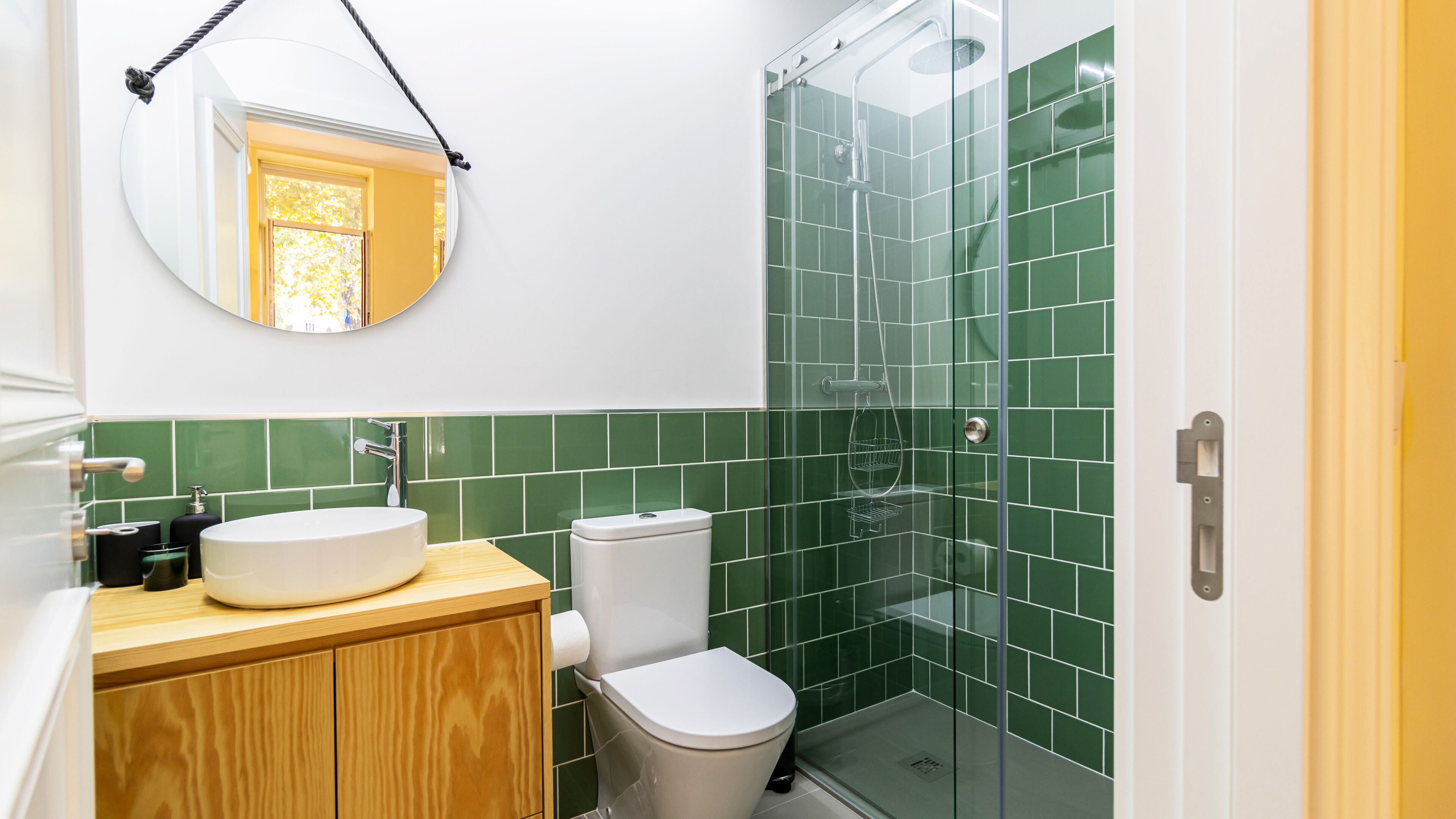 A bathroom with a wooden vanity, a white sink, a toilet, and a glass shower enclosure with green tiles.