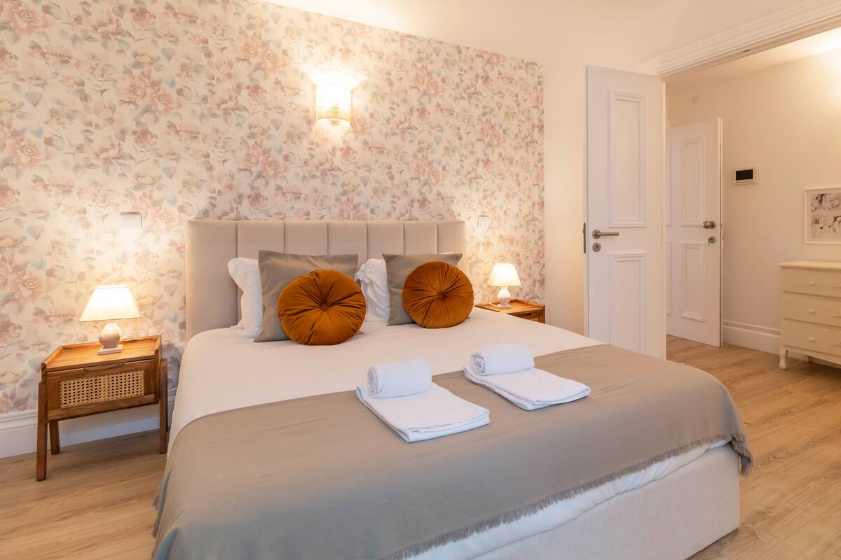 A neatly made bed with a grey bedspread, two brown pillows, and two rolled towels. There are two bedside tables with lamps and a wall-mounted light fixture. The room has floral wallpaper and wooden flooring.