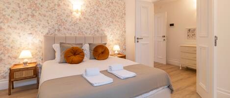 A neatly made bed with a grey bedspread, two brown pillows, and two rolled towels. There are two bedside tables with lamps and a wall-mounted light fixture. The room has floral wallpaper and wooden flooring.