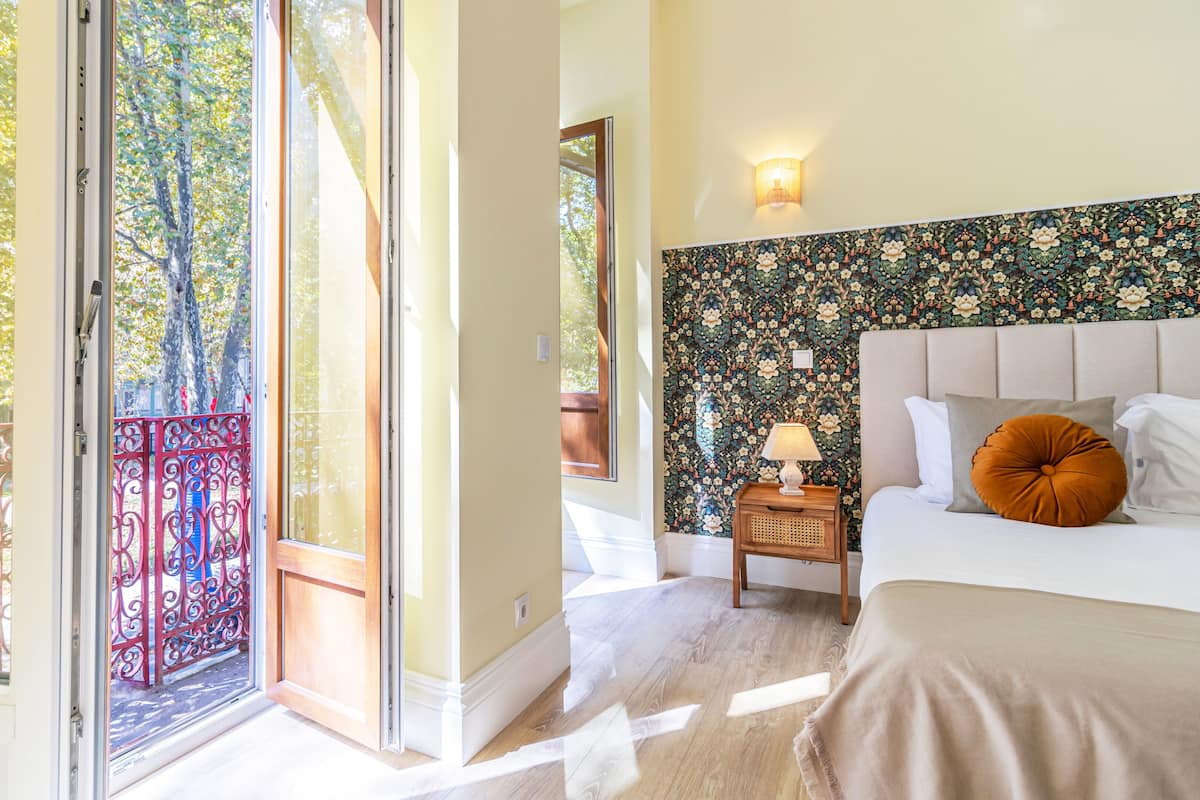 A bedroom with a bed, a nightstand with a lamp, a window with a view of trees, and a patterned wall.