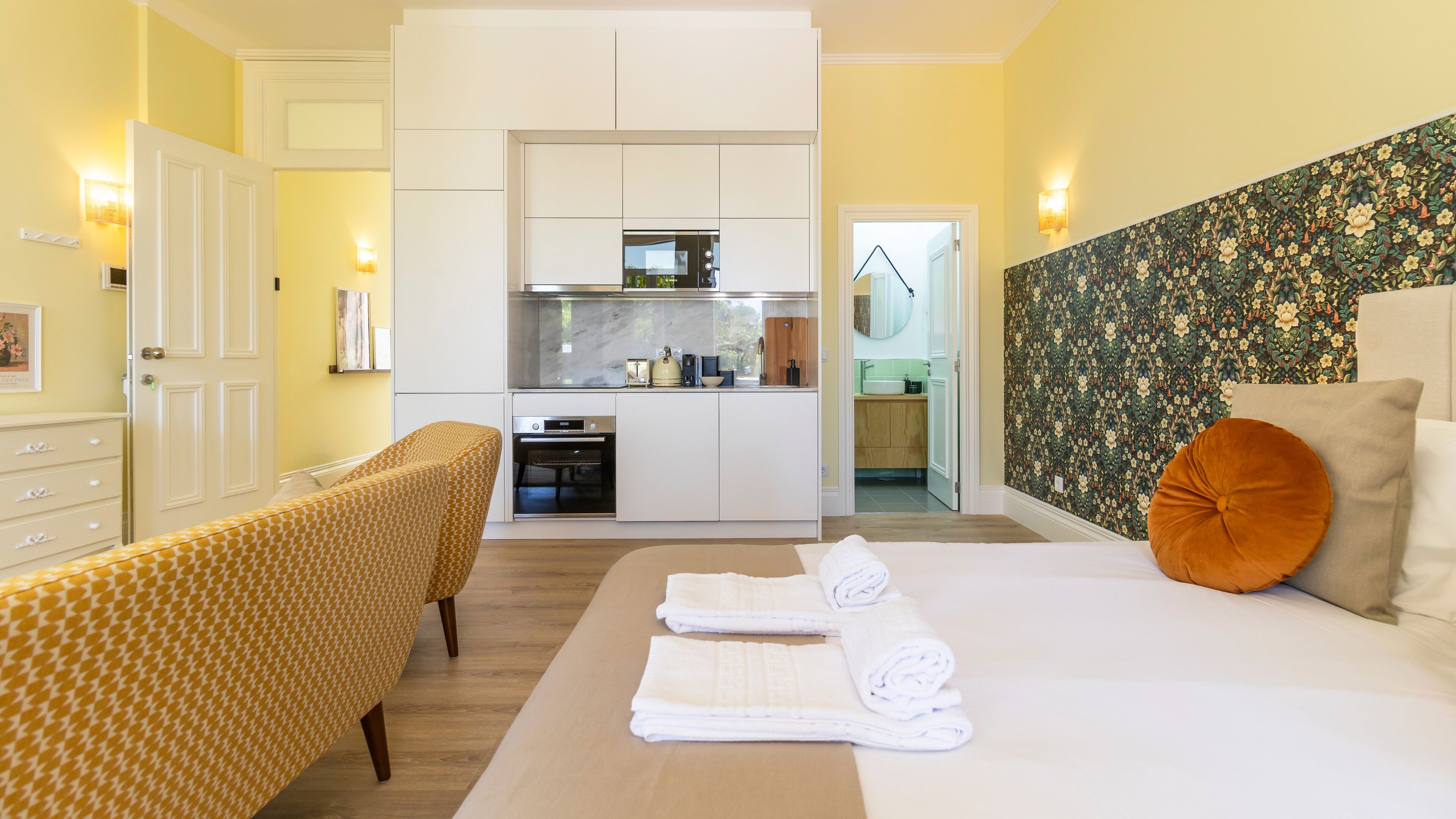 A hotel room with a bed, a sofa, a kitchen area, and a bathroom visible through an open door.