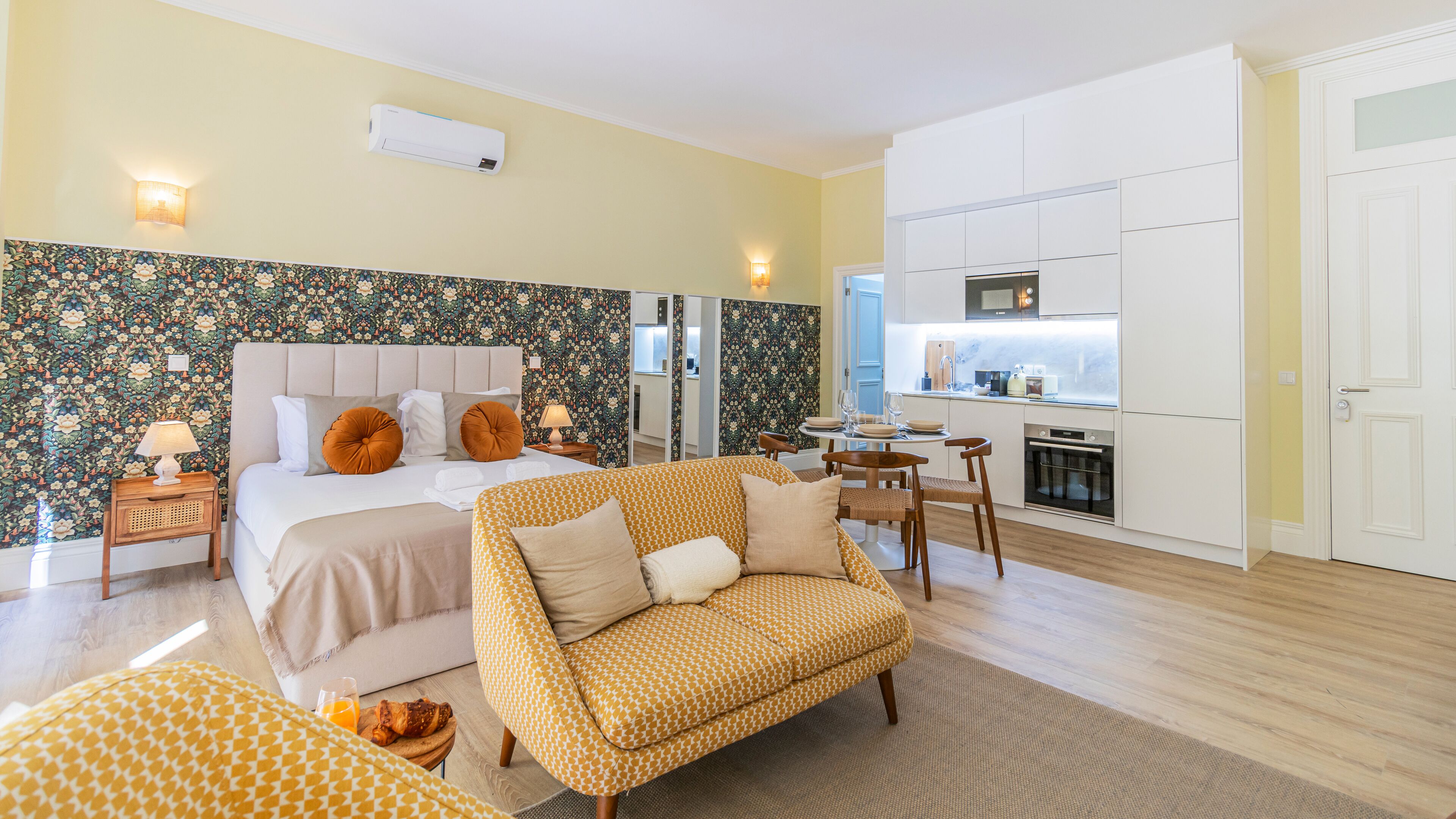 A modern hotel room with a bed, a sofa, a dining table, and a kitchen area.