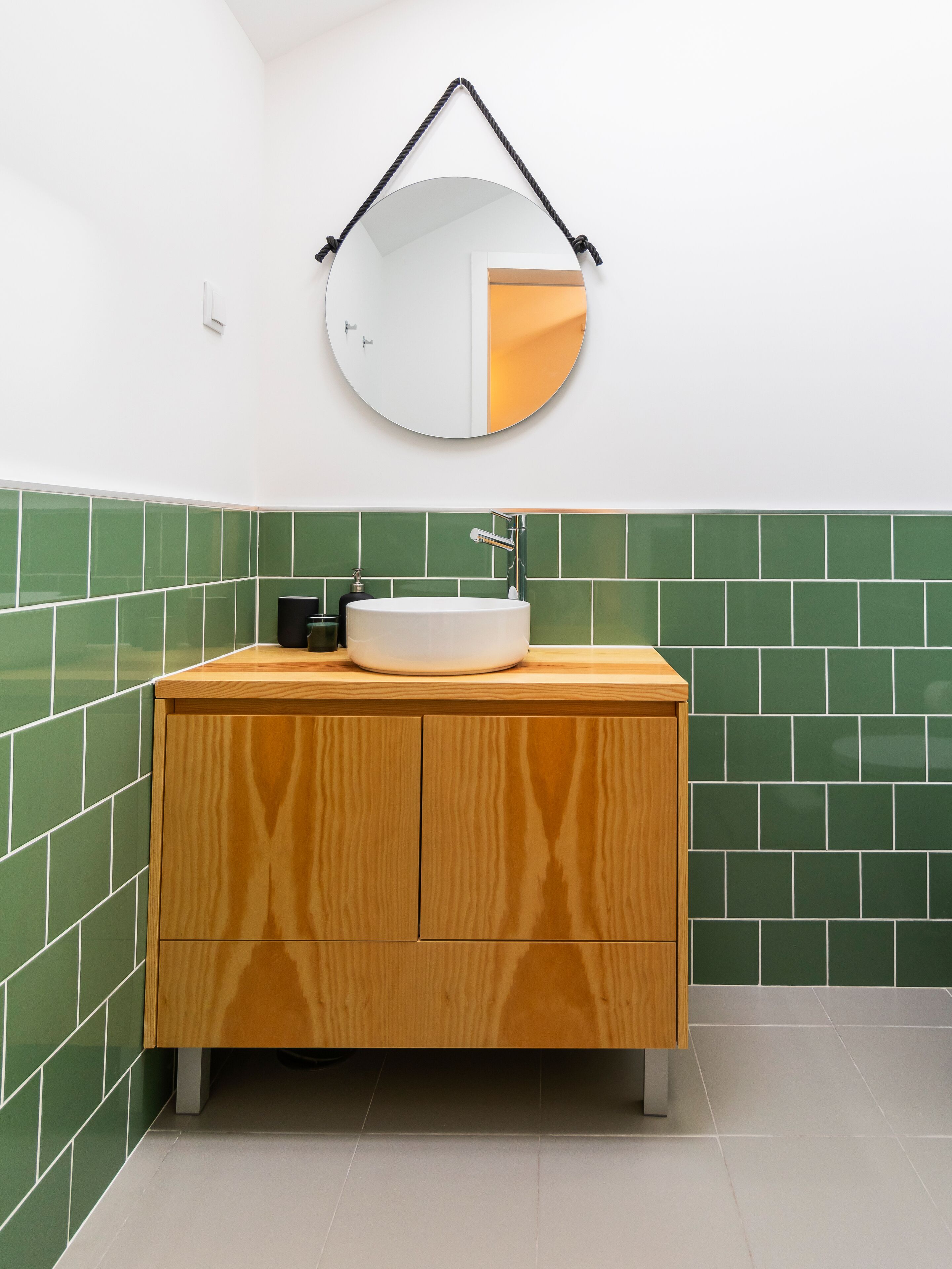 A bathroom with a wooden vanity, a white sink, and green tiled walls.