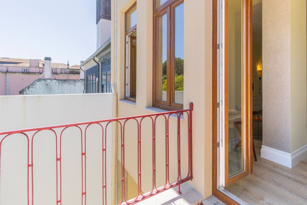 A balcony with a red metal railing, a sliding glass door leading to a room with a wooden floor, and a view of neighboring buildings.