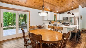 A spacious dining area with a large wooden table and chairs, a kitchen with white cabinets and stainless steel appliances, and a view of a wooded area through glass doors. - Blueberry: 3  BR, 3  BA House in Canaan Valley, Sleeps 6 (Canaan Valley)