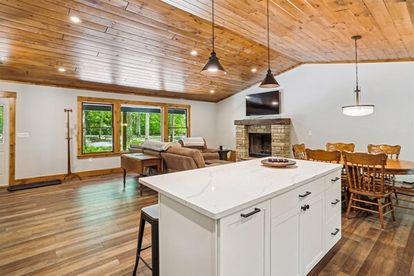 A spacious living area with a large wooden ceiling, a stone fireplace, a white kitchen island, and a dining area with wooden chairs.