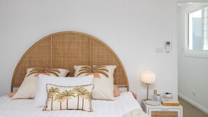A bed with a woven headboard, palm tree pillows, a bedside lamp, and a small bedside table with a book and a candle. - Bohemian Coastal Dream (Port Macquarie)