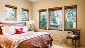 A bedroom with a bed, a chair, and large windows with a view of trees.