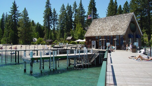 A wooden dock extending into clear water, surrounded by trees. A building with a sloped roof and large windows is visible.