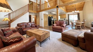 A spacious living area with leather sofas, a wooden coffee table, and a dining area with wooden chairs. A staircase leads to an upper level.