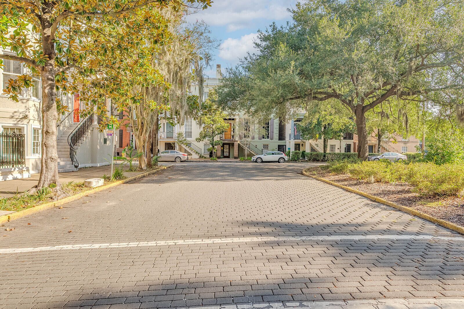 A paved street lined with trees and parked cars, leading to a building with a staircase and a flag.