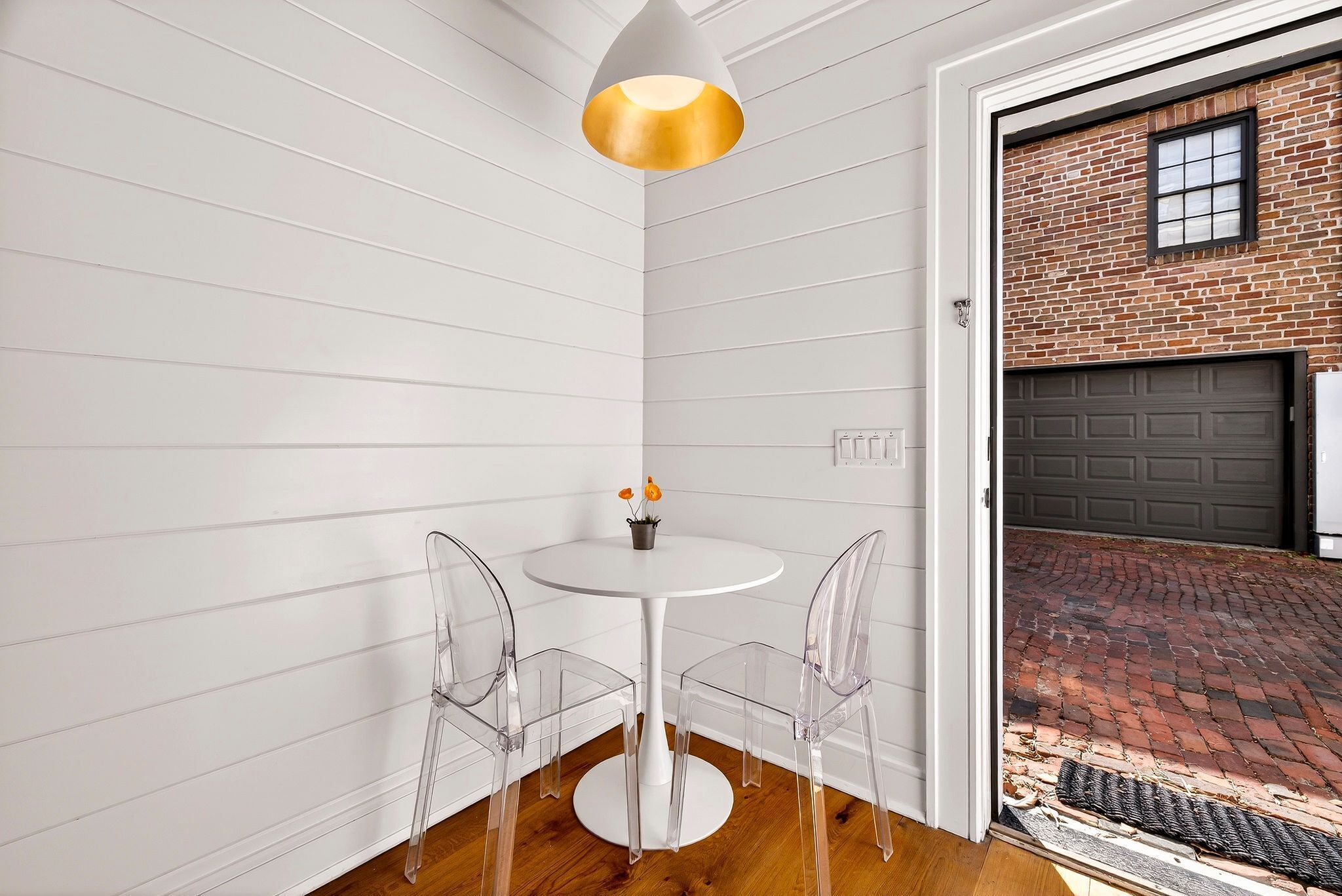 A corner room with a white paneled wall, a round white table with two clear chairs, a gold pendant light, and a view of a brick driveway through an open door.