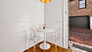 A corner room with a white paneled wall, a round white table with two clear chairs, a gold pendant light, and a view of a brick driveway through an open door. - Newly Built Carriage House in Unbeatable Location by Lucky Savannah (Savannah)
