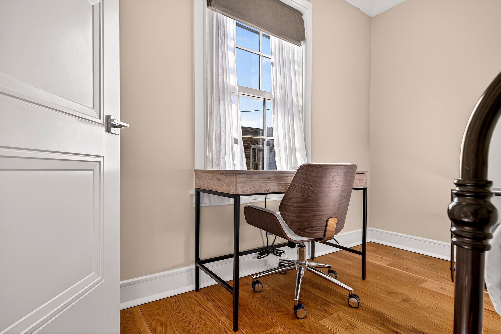 Une pièce avec un bureau en bois, un fauteuil de bureau pivotant, une fenêtre avec des rideaux blancs et une porte.