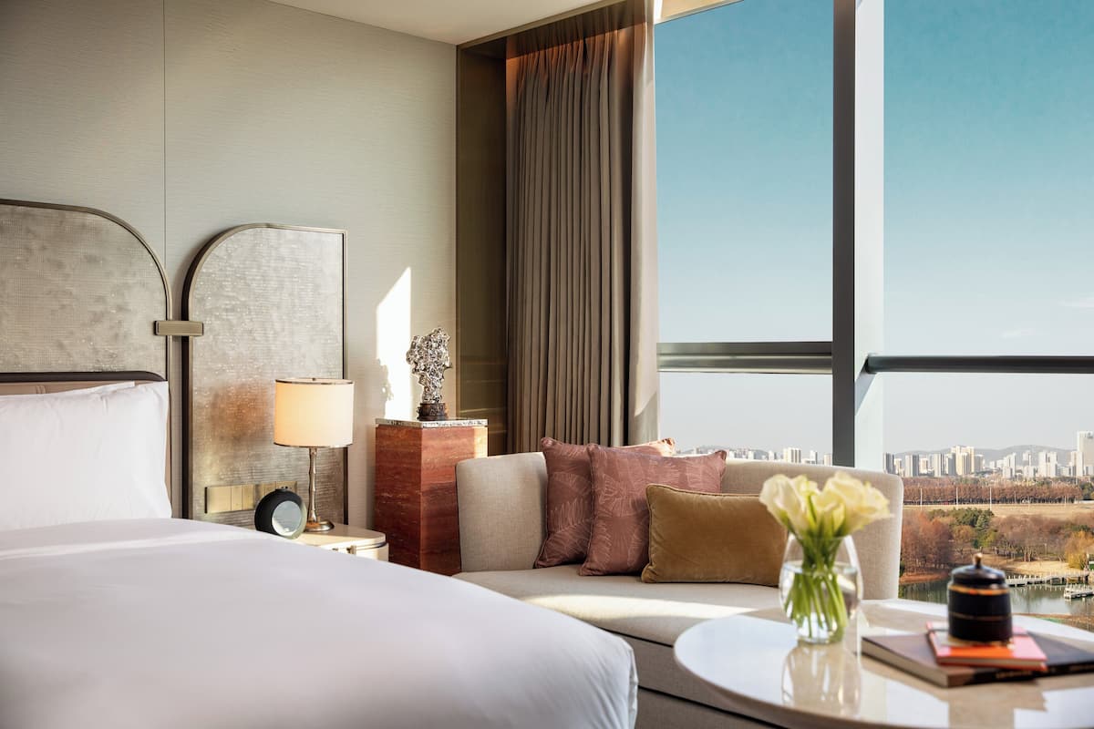 Een moderne hotelkamer met een groot bed, een bank, een kleine tafel met een bloevas en uitzicht op een stadsgezicht door grote ramen.