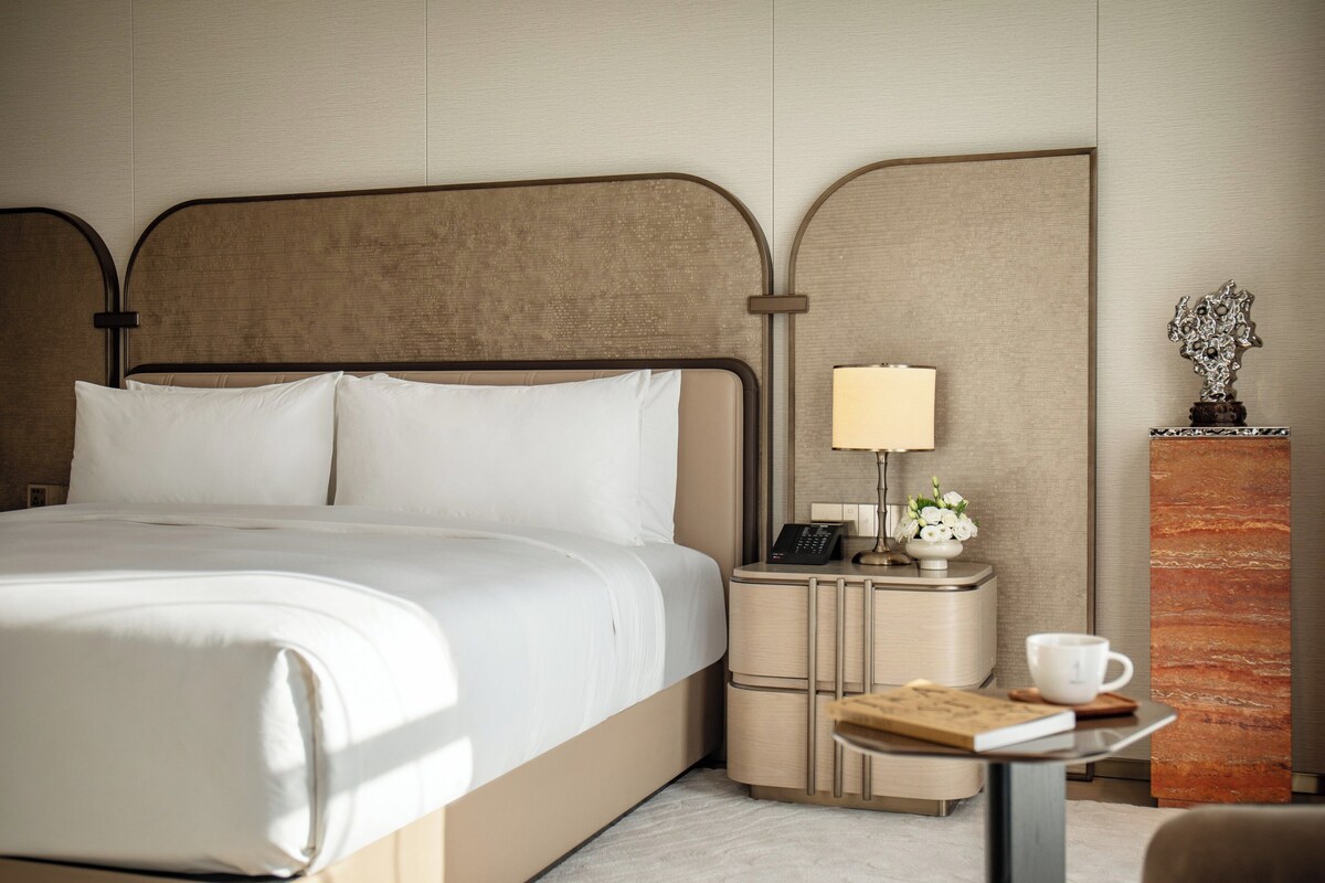 Een hotelkamer met een groot bed, nachtkastjes, een lamp, een telefoon, een klein tafeltje met een boek en een kopje, en een decoratief object aan de muur.