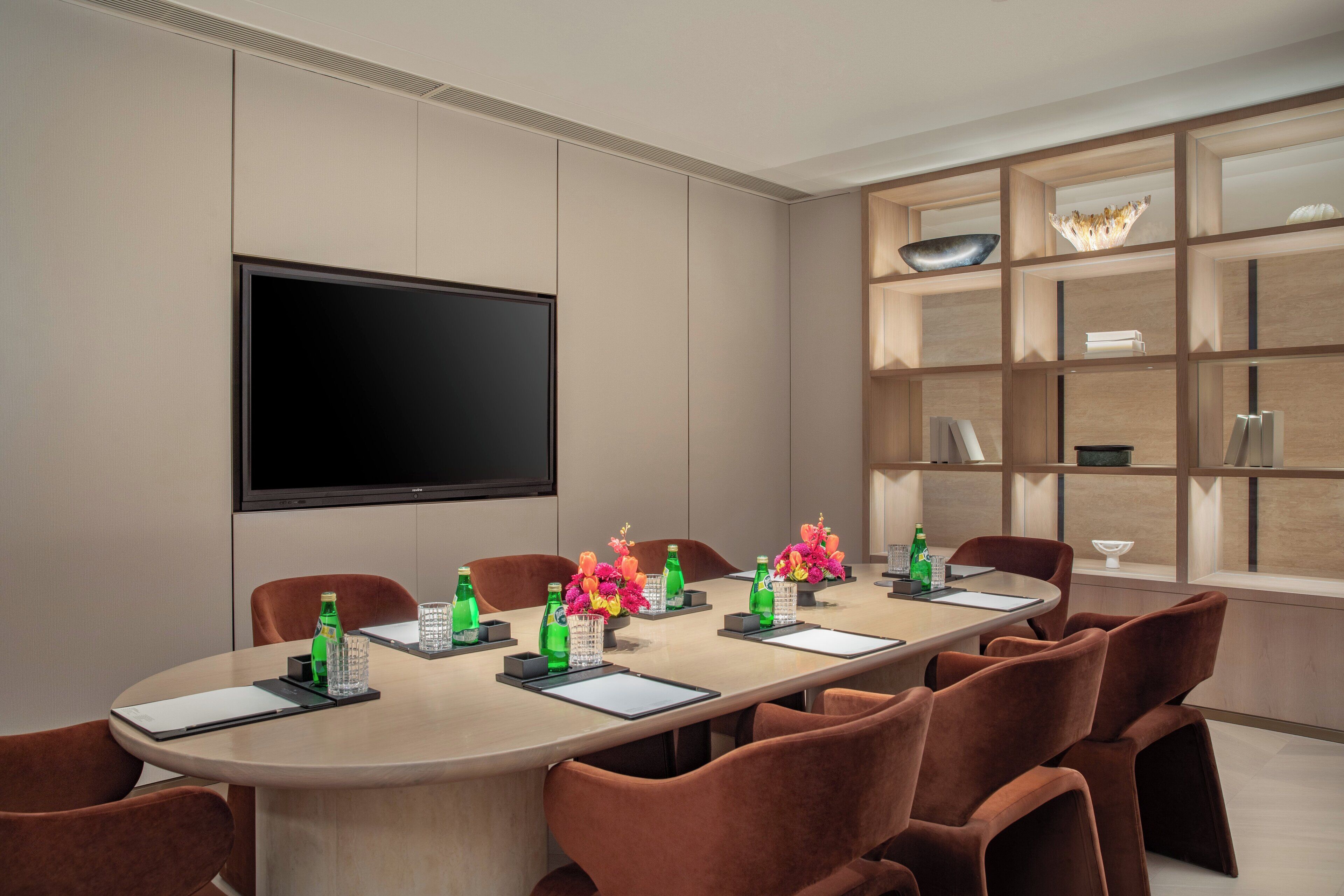 A conference room with a long table, brown chairs, a flat-screen TV, and a bookshelf with decorative items.