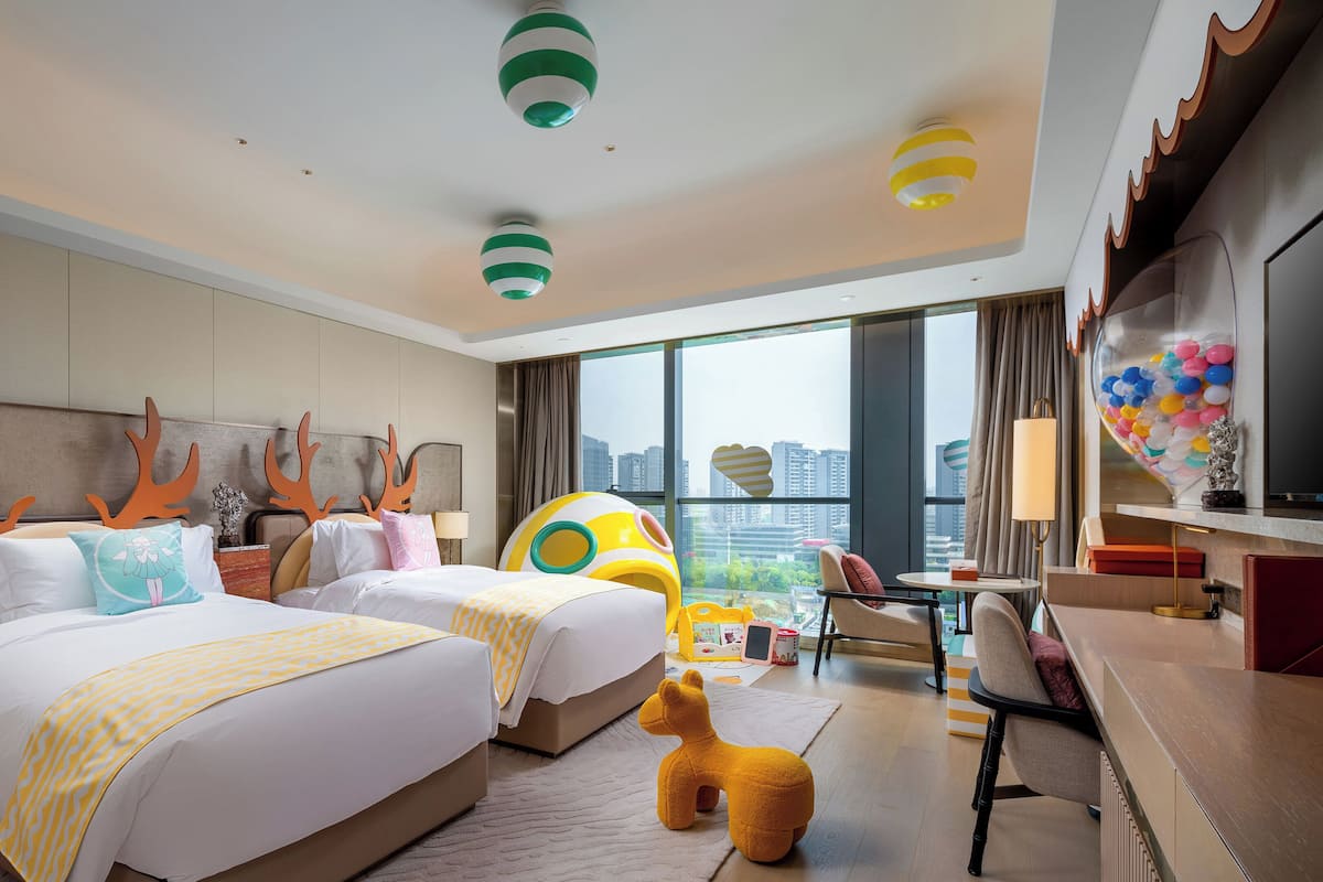 Hotelkamer met twee bedden, een bureau, een stoel, een teddybeer en een groot raam met uitzicht op de stad.