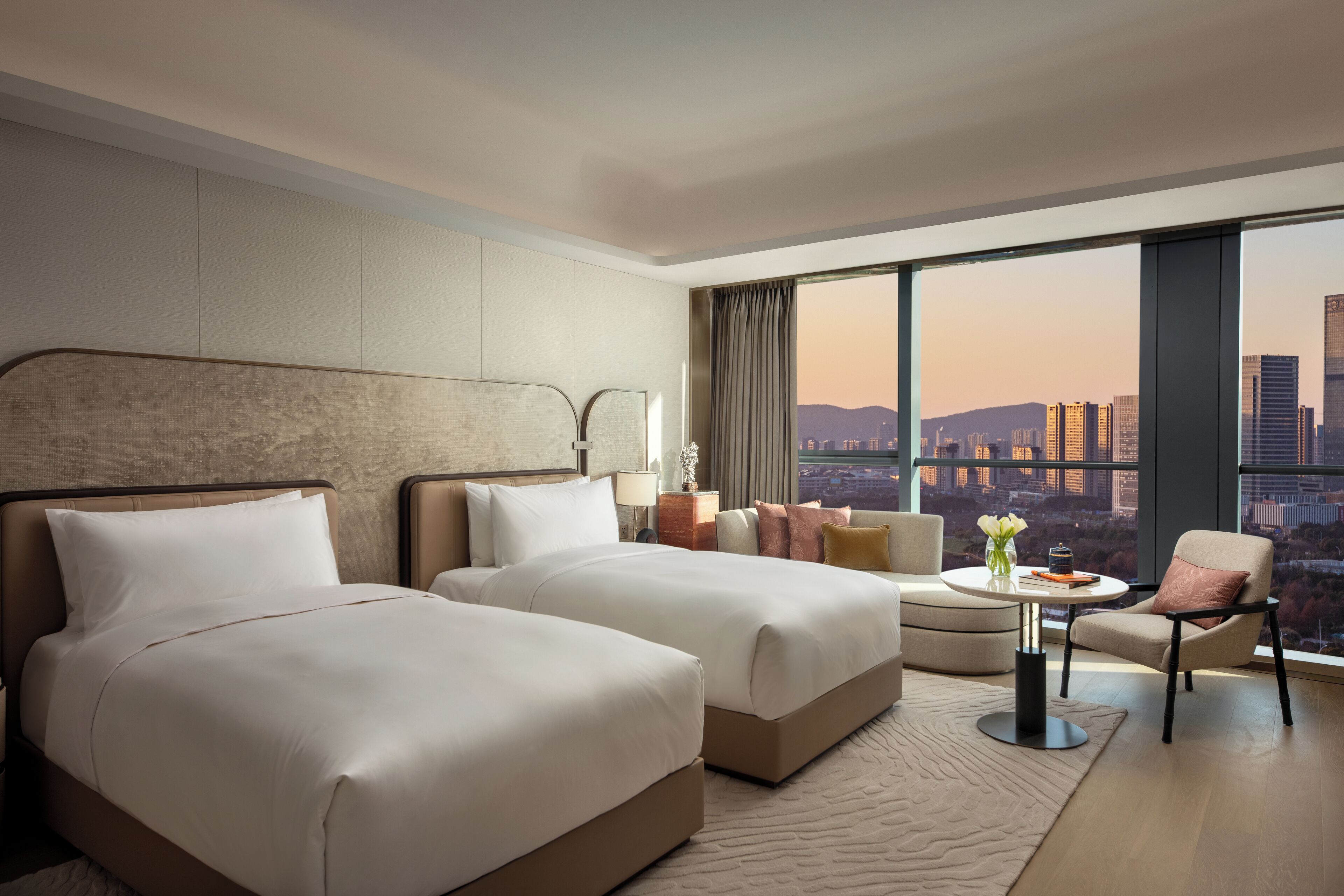 A hotel room with two beds, a sofa, a small table, and a chair. The room has a large window with a city view.