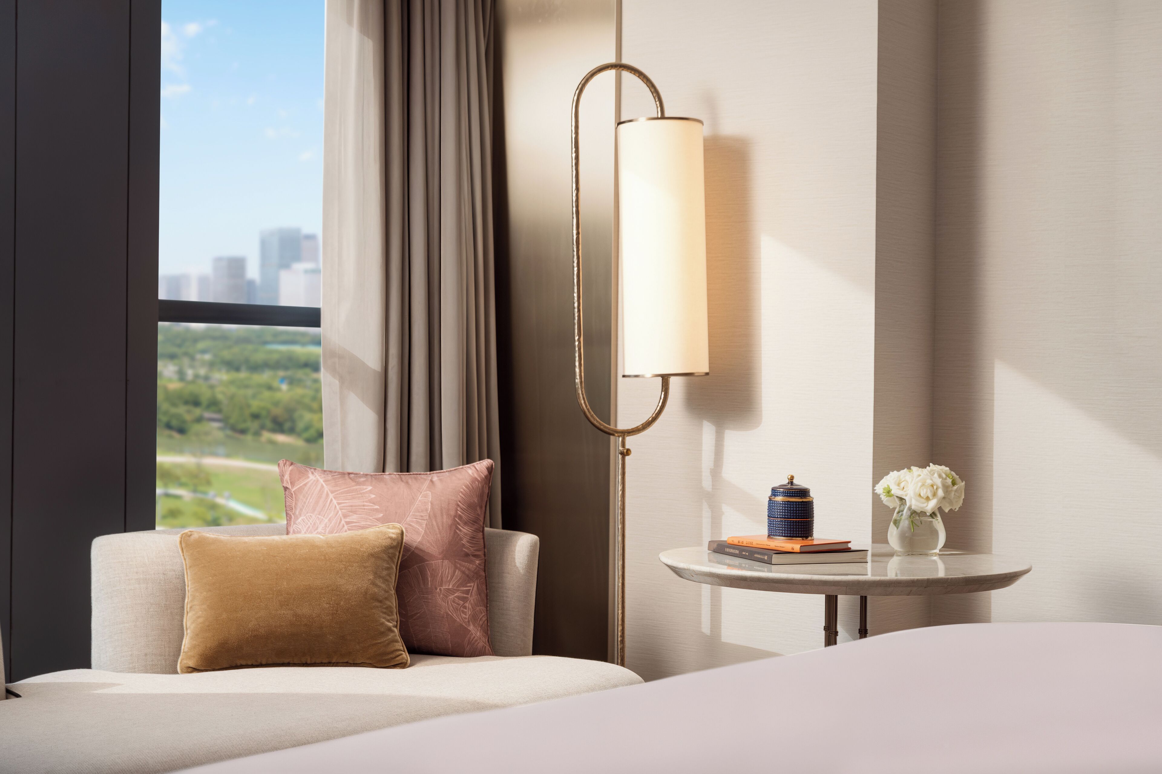 Habitación de hotel con cama, mesita de noche con lámpara, ventana con vistas a zonas verdes y edificios, y una pequeña mesa con un jarrón de flores.
