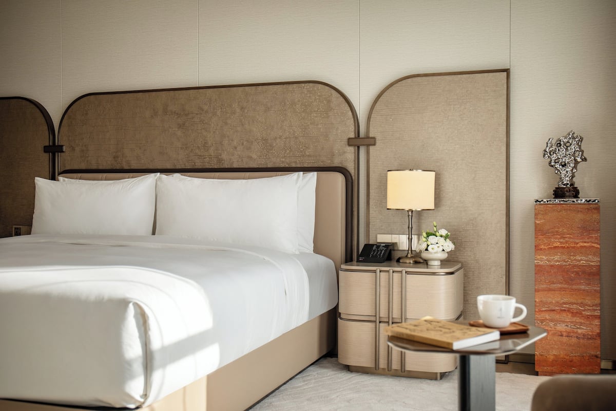 Een hotelkamer met een groot bed, nachtkastje met een lamp, een klein tafeltje met een kopje en een boek, en een decoratief object aan de muur.