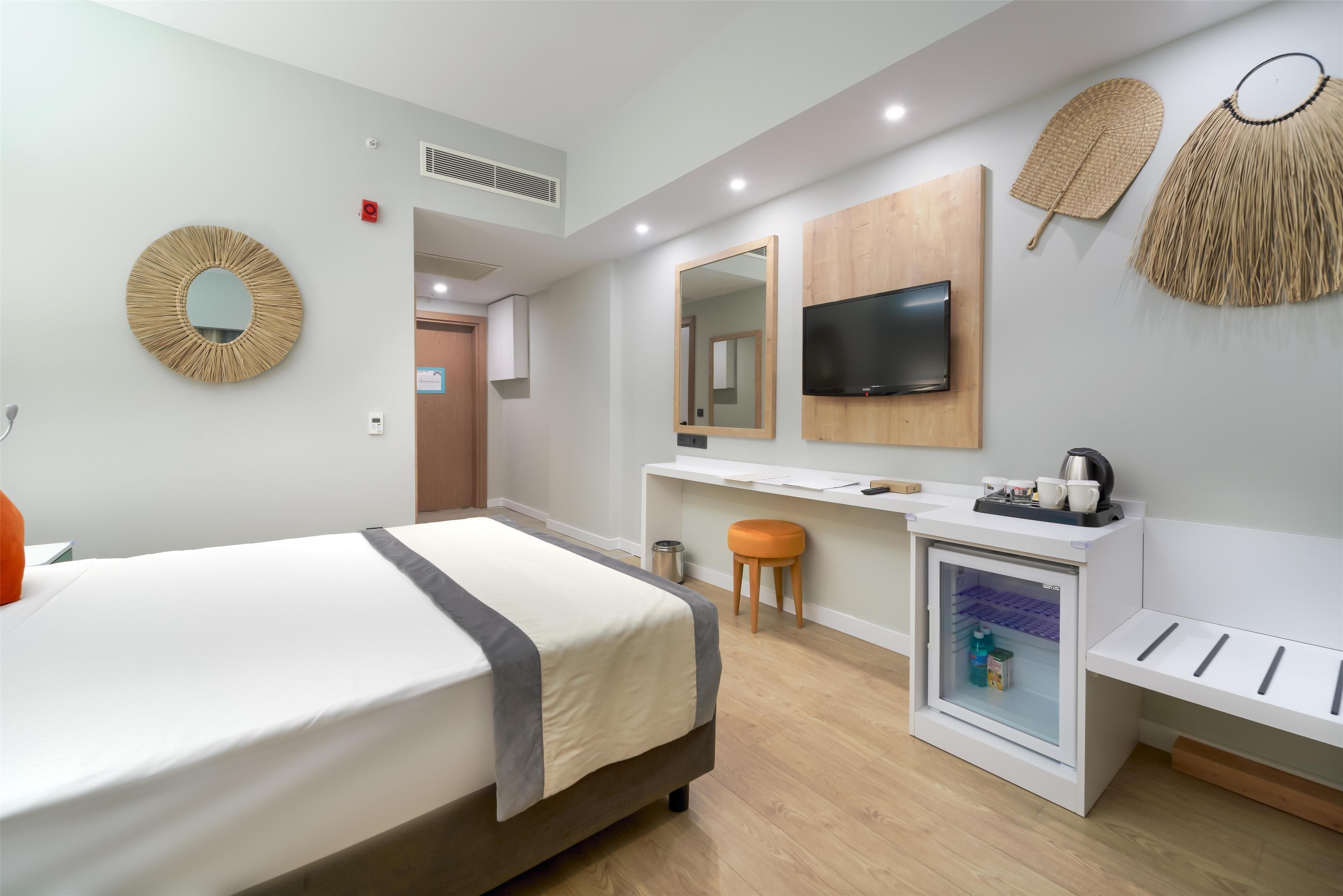 A hotel room with a bed, a desk with a TV, a minibar, and a mirror.