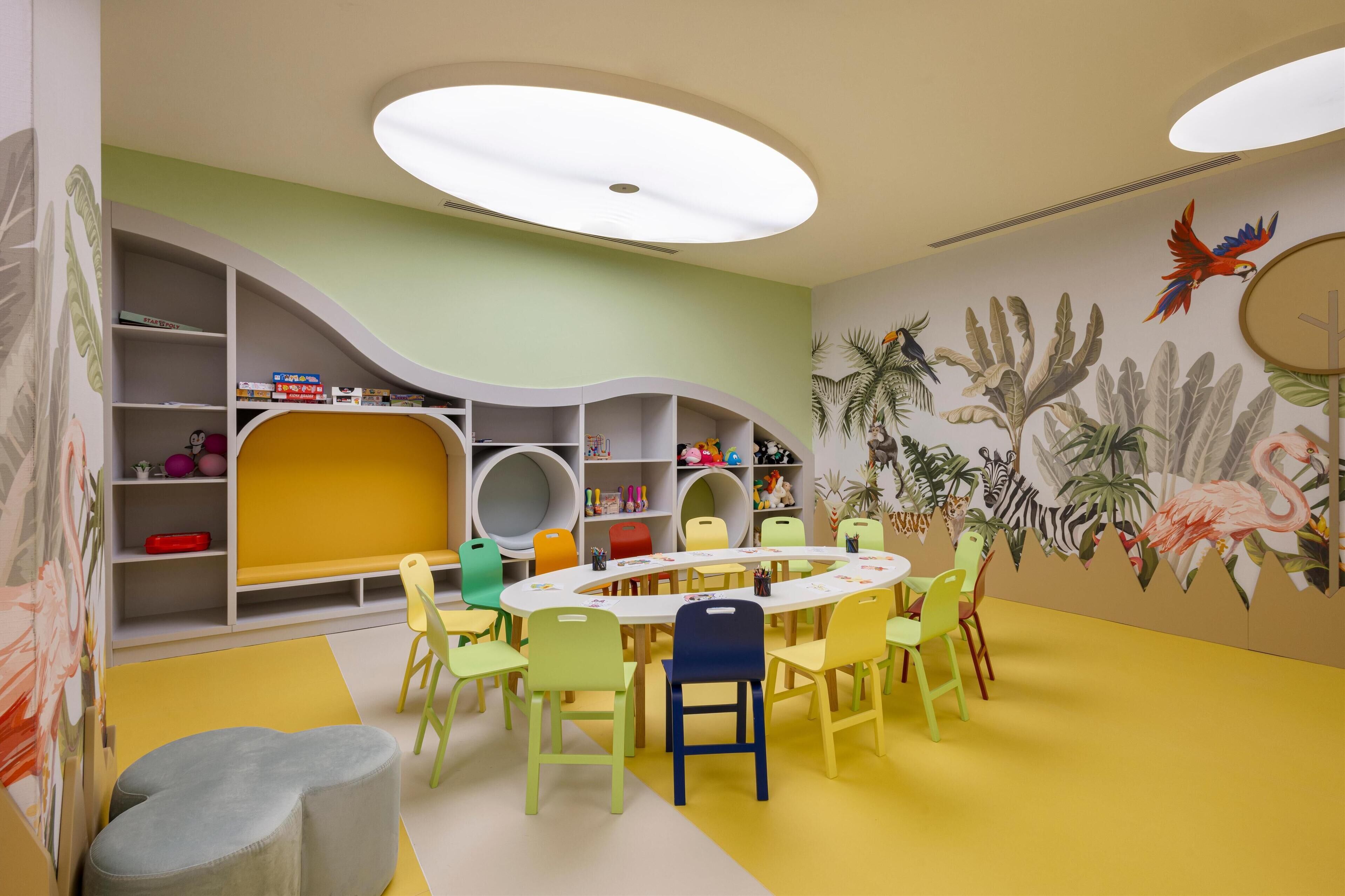 Una sala de juegos infantil con una mesa redonda, sillas de colores y un mural de animales en la pared.