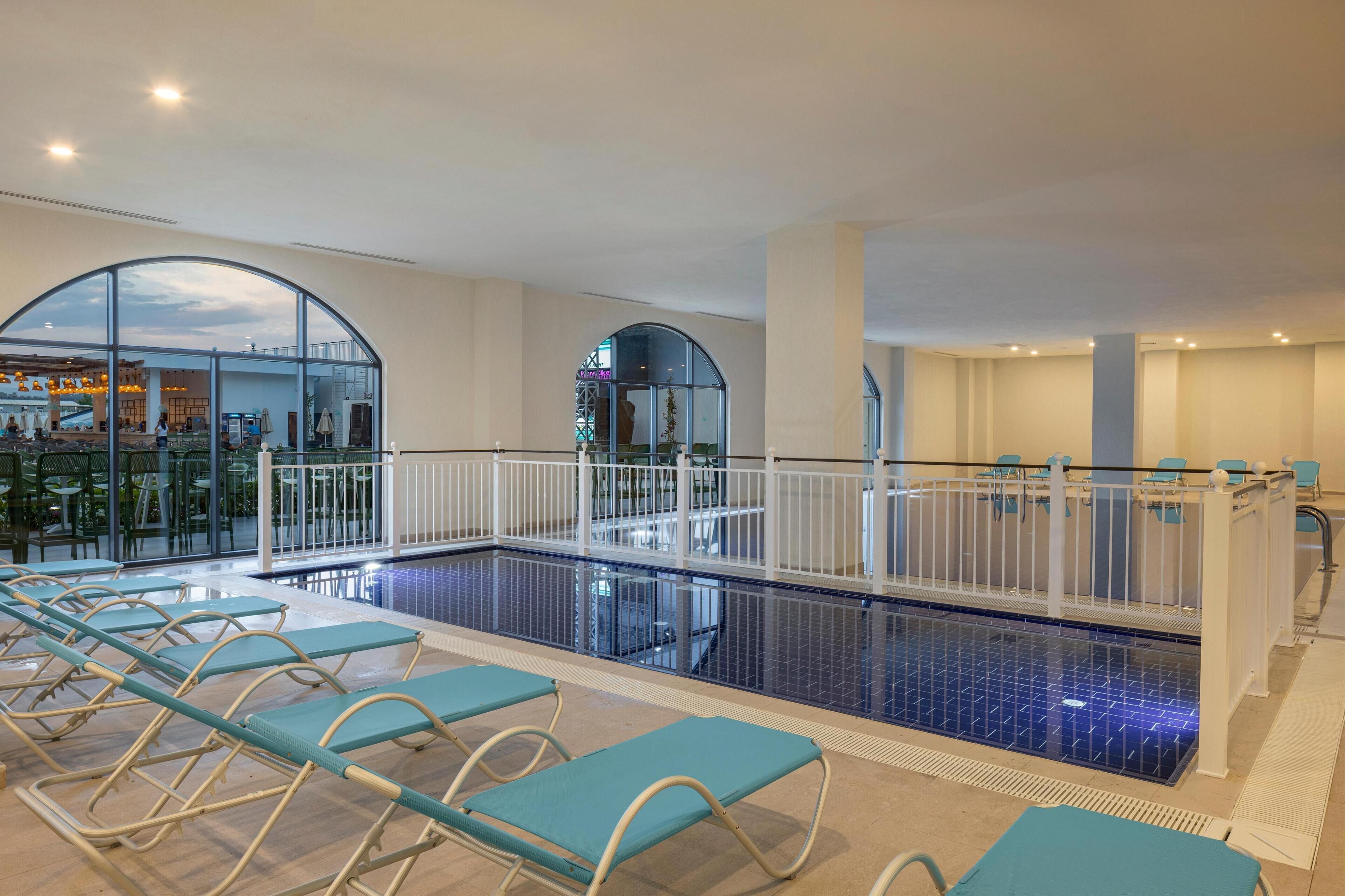 Una piscina interior con sillones de descanso, barandilla de cristal y vista panorámica de la ciudad a través de amplios ventanales.