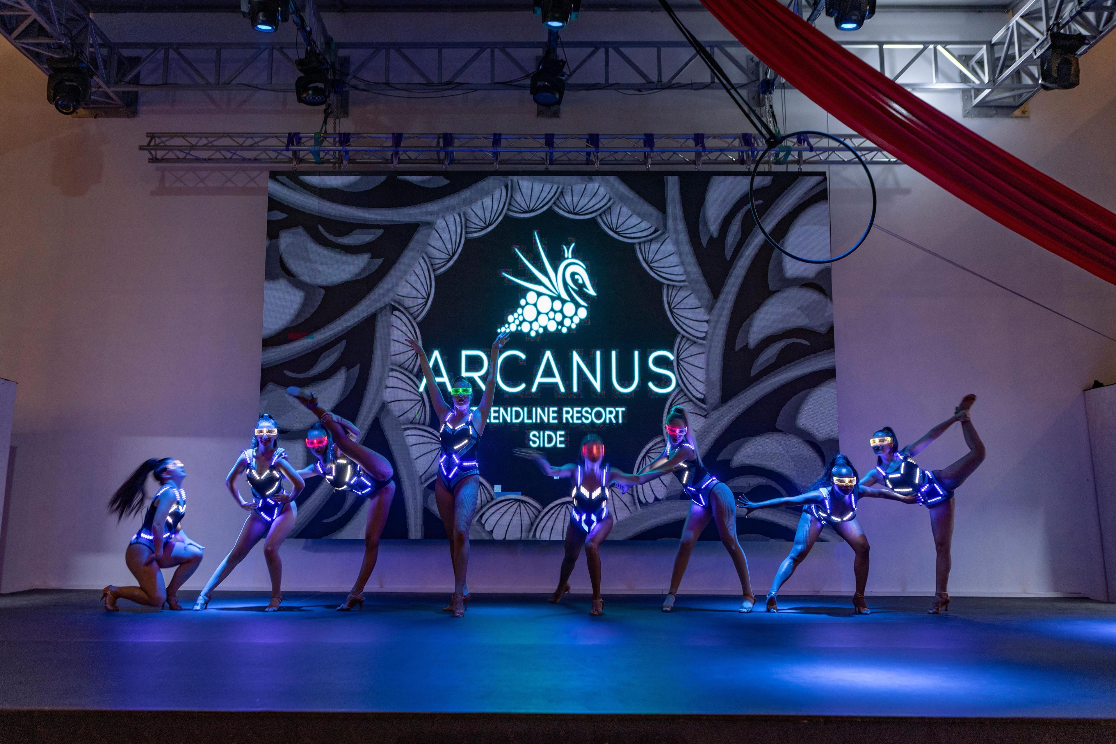 Un escenario con bailarines con vestuarios iluminados y un fondo con el texto "ARCANUS ENDLINE RESORT SIDE".