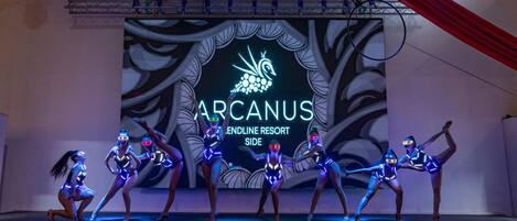 Un escenario con bailarines con vestuarios iluminados y un fondo con el texto "ARCANUS ENDLINE RESORT SIDE".